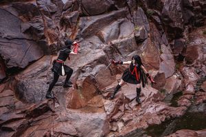 Two cosplayers in black costumes with red details leap and clash with swords on a steep red rock wall.