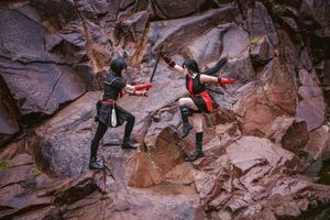 Two cosplayers in black and red costumes duel with swords mid-action on a rugged red rock cliff.