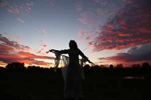 Silhouette of a woman in a sheer flowing cape with outstretched arms against a vivid pink and purple sunset sky.