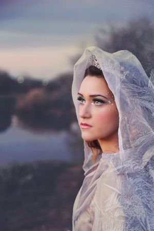 Portrait of a woman in a frosted sheer veil and jeweled tiara with sparkling makeup, set against a soft misty lake.