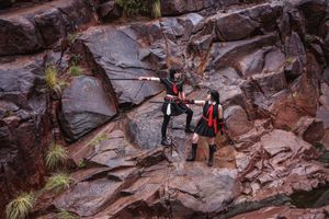 Two cosplayers in black and red costumes aim their swords at each other on a flat rock among desert plants.