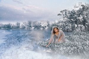 Woman in a sheer hooded gown kneeling beside a frozen lake in a snowy winter landscape with frost-covered trees.