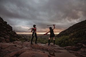 Two cosplayers raise swords in a dramatic duel on rocky ground beneath a stormy sunset sky.