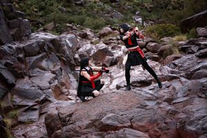 Two cosplayers in black and red outfits with weapons pose on rugged rocky terrain in the desert.