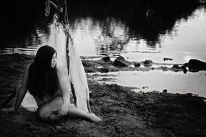 Black and white photo of a woman with long dark hair sitting on a rocky lakeshore beside draped white fabric.