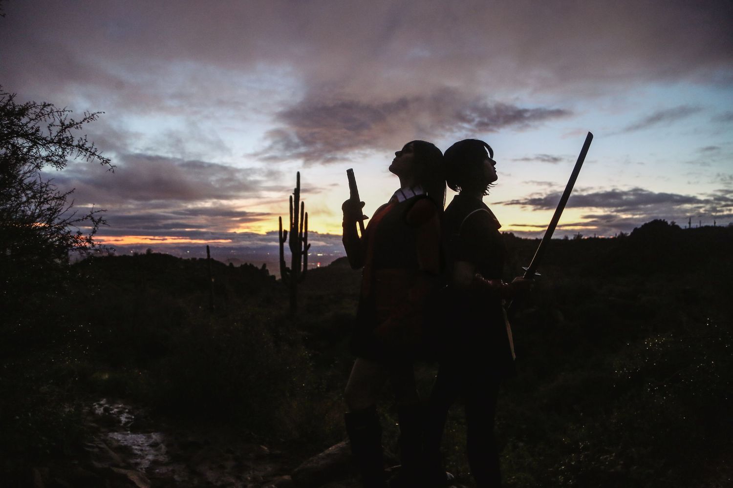 Silhouettes of two cosplayers holding weapons stand among saguaro cacti against a moody desert sunset sky.