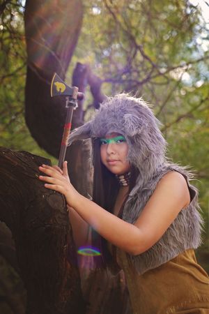 Young woman in a gray fur hood and fur vest holds a small axe beside a tree trunk, with sunlight filtering through the leaves.