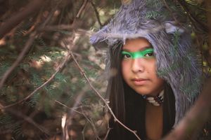 Young woman with bright green face paint peers through evergreen branches wearing a gray wolf fur hood in a forest.