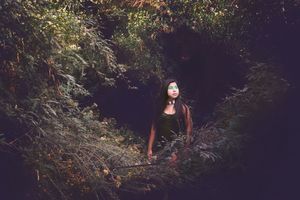 Woman with glowing green eye makeup stands in a dark, mossy forest clearing surrounded by dense foliage.