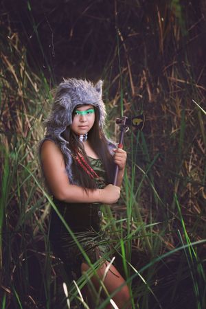 Woman in a gray wolf fur hood and green eye paint holds a small hatchet while standing in tall marsh grass.
