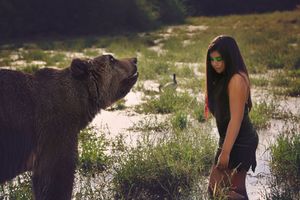 Woman with long dark hair stands in a wetland meadow facing a large brown bear with its head raised.
