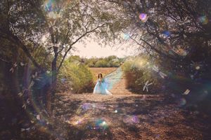 Woman in a flowing pale blue gown walks along a sunlit path under trees with dreamy lens flares and bokeh light.