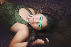 Woman in a green dress lies on the forest floor with green face paint, a beaded collar and a feather in her hair.