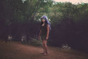 Girl with silvery-blue hair stands on a dirt path in the woods as several raccoons gather around her at dusk.