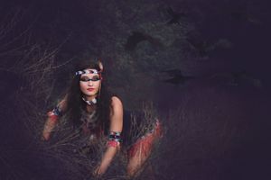 Woman with dark eye paint and a beaded headband crouches in dry brush wearing red-tasseled armbands in dim moody light.