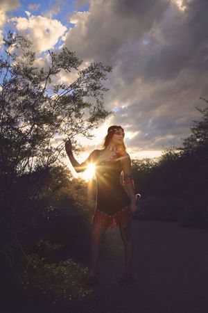 Silhouetted woman in beaded headband and fringed skirt raises her arm as the setting sun bursts through clouds on a trail.
