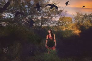 Woman in beaded tribal-style attire stands in a dusky forest clearing as black birds fly overhead at sunset.