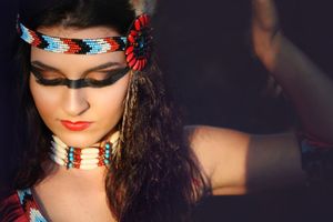 Close-up portrait of a woman wearing a beaded headband, feather ornament and layered beaded choker necklace in warm light.