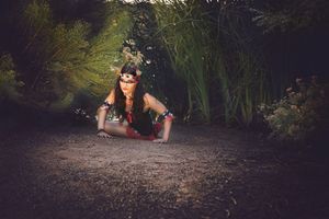 Woman in beaded headband crouches low on a dirt path beside tall green reeds in a moody, dimly lit woodland scene.