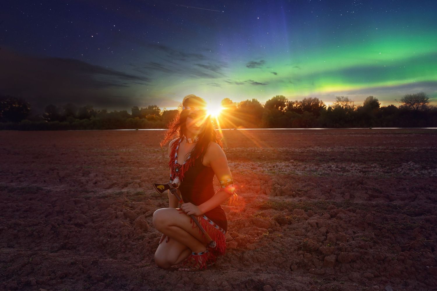 Woman kneels in an open field at twilight beneath a glowing sunset and green northern lights in a starry sky.