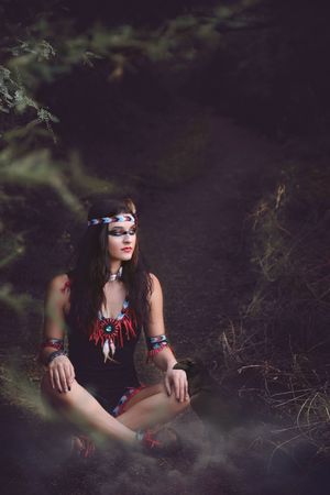 Woman with beaded headband and feather necklace sits cross-legged on a misty forest trail in soft, moody light.