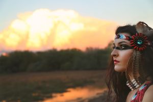 Side profile of a woman with beaded headband and feather earrings beside a river as glowing sunset clouds fill the sky.