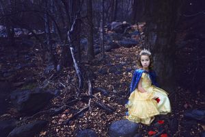 Girl in a yellow princess dress, blue cape and tiara sits on forest rocks among bare trees, fallen leaves and red apples.