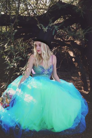 Blonde woman in a black top hat and a flowing teal tulle ball gown sits beneath a twisted tree in a whimsical woodland portrait.
