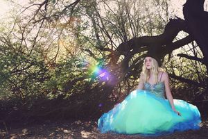 Woman in a turquoise tulle gown sits under gnarled tree branches as colorful sunlight flares create a dreamy, fairytale forest scene.