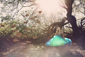 Woman in a green and blue ball gown sits in a sunlit forest while book pages float through the air in a magical, storybook setting.