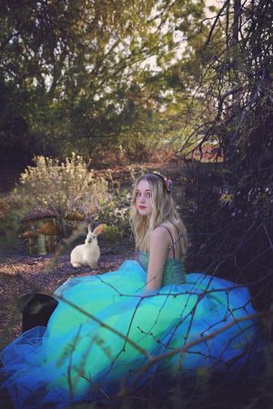 Woman in a flowing blue-green gown with a flower in her hair sits in a woods beside a white rabbit and mushrooms in a fairytale scene.