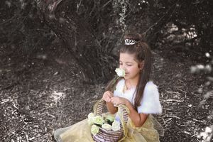 Young girl in a tiara, white fur wrap and yellow tulle dress smells a white rose while holding a basket of flowers in a shaded woodland.