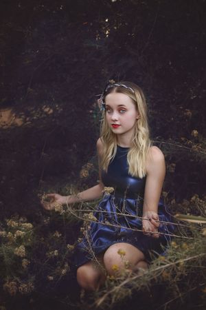 Young woman with blonde hair and red lipstick sits among dried wildflowers in a shiny navy dress in a moody forest.