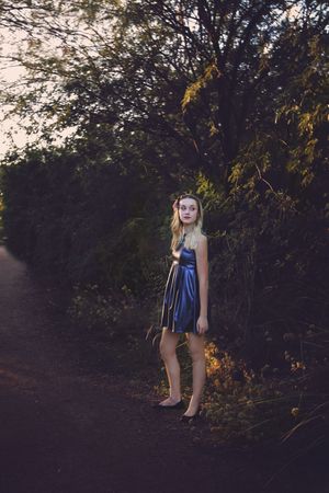 Blonde young woman in a metallic blue dress stands on a shaded dirt trail beside lush green trees at sunset.