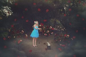 Girl in a blue dress stands in a dark enchanted garden with falling red rose petals, flowers and a small white rabbit nearby.