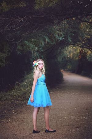 Young woman in a turquoise tulle dress with a colorful hair flower stands on a tree-lined dirt path.