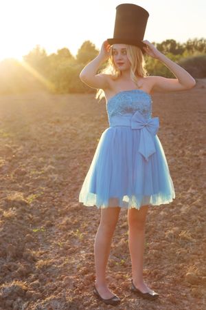 Woman in a pale blue tulle dress with a bow holds a tall black top hat in a sunlit field at golden hour.