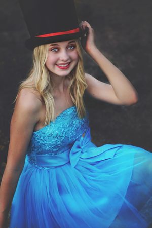 Smiling blonde woman in a sparkly blue dress tips a black top hat with a red band in a playful portrait.
