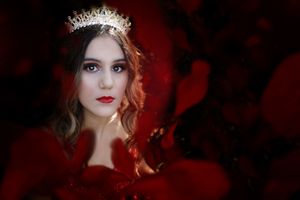 Young woman wearing a jeweled tiara and red lipstick poses in a dramatic portrait surrounded by deep red tones.