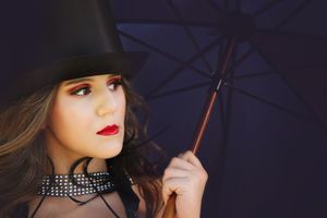 Close-up portrait of a woman in a black top hat, red lipstick and studded choker holding a dark umbrella.