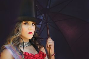 Woman in a black top hat, rhinestone choker and red ruffled top holds a dark umbrella in a moody theatrical portrait.