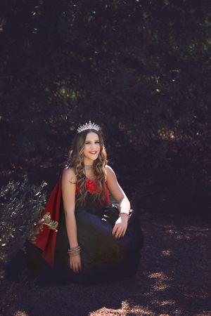 Young woman in a tiara and black gown with red cape and red floral accent sits in a sunlit wooded setting for a fairytale portrait.