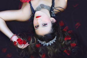 Woman lying back with a jeweled necklace, red lipstick and red bracelet, surrounded by scattered red rose petals on a dark backdrop.