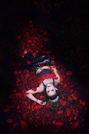 Overhead view of a woman in a tiara and dark dress lying amid a bed of red roses and petals in a dramatic dark setting.