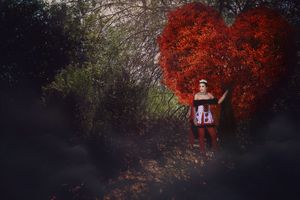 Queen of hearts costume portrait with a woman standing before a giant red heart-shaped bush on a misty forest path.