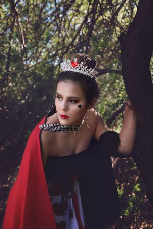 Woman dressed as a playing card queen of hearts with a sparkling crown, red cape and heart face paint, leaning on a tree in the woods.