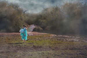 Small child in a teal dress with sheer fairy wings stands in a misty meadow with trees in the background, creating a magical scene.