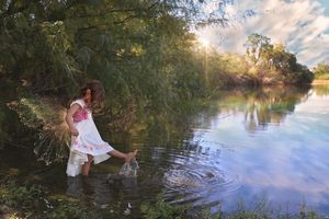 Girl in a white dress with pink flowers wades through a calm lake, splashing the water under green trees at sunset.