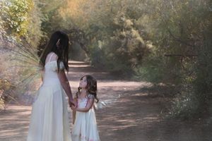 Woman in a long cream gown holds the hand of a little girl in a fairy dress and wings on a sunlit forest path.