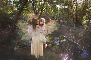 Woman in a cream gown with large translucent fairy wings holds a child beside a woodland stream with dragonflies and trees.